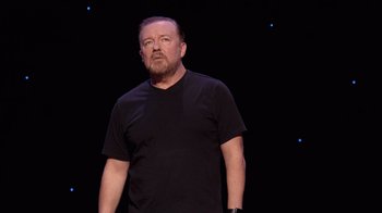 Movie still from “Ricky Gervais: SuperNature” (2022), directed by John L. Spencer – A man in a black t - shirt stands in front of a black background; Medium shot, Low angle
