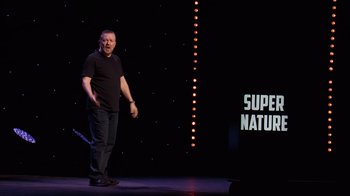 Movie still from “Ricky Gervais: SuperNature” (2022), directed by John L. Spencer – A man standing on a stage in front of a microphone; Wide shot, Over the shoulder angle