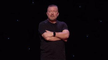 Movie still from “Ricky Gervais: SuperNature” (2022), directed by John L. Spencer – A man standing in front of a black background; Medium shot, High angle