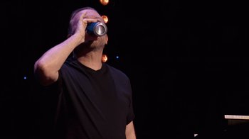 Movie still from “Ricky Gervais: SuperNature” (2022), directed by John L. Spencer – A man holding a can of beer up to his face; Medium shot, Low angle