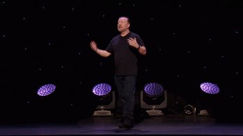Movie still from “Ricky Gervais: SuperNature” (2022), directed by John L. Spencer – A man standing on a stage in front of a crowd; Wide shot, Low angle