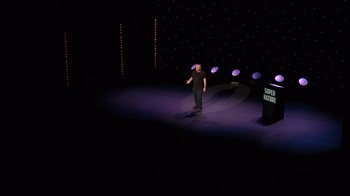 Movie still from “Ricky Gervais: SuperNature” (2022), directed by John L. Spencer – A man standing on a stage in front of a microphone; Extreme Wide shot, High angle