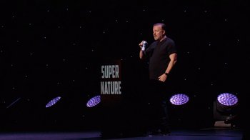 Movie still from “Ricky Gervais: SuperNature” (2022), directed by John L. Spencer – A man standing in front of a microphone on a stage; Medium shot, Over the shoulder angle