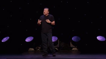 Movie still from “Ricky Gervais: SuperNature” (2022), directed by John L. Spencer – A man standing on a stage in front of purple lights; Medium shot, Low angle