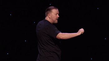 Movie still from “Ricky Gervais: SuperNature” (2022), directed by John L. Spencer – An image of a man that is standing up on stage; Medium shot, High angle