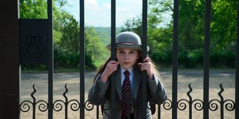 Movie still from “Roald Dahl's Matilda the Musical” (2022), directed by Matthew Warchus – A girl in a suit and tie standing in front of an iron fence; Medium shot, High angle
