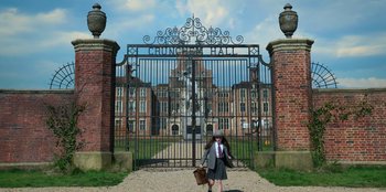 Movie still from “Roald Dahl's Matilda the Musical” (2022), directed by Matthew Warchus – A woman in a suit and tie walking in front of an iron gate; Wide shot, Low angle