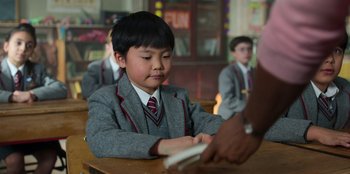 Movie still from “Roald Dahl's Matilda the Musical” (2022), directed by Matthew Warchus – A young boy in a suit and tie is playing a video game; Close Up shot, Over the shoulder angle