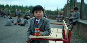 Movie still from “Roald Dahl's Matilda the Musical” (2022), directed by Matthew Warchus – A boy in a school uniform sitting on a bench; Medium shot, High angle