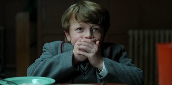 Movie still from “Roald Dahl's Matilda the Musical” (2022), directed by Matthew Warchus – A young boy sitting at a table with his hands over his mouth; Close Up shot, High angle