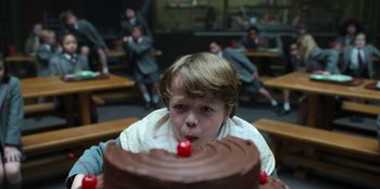 Movie still from “Roald Dahl's Matilda the Musical” (2022), directed by Matthew Warchus – A boy blowing out a candle on a chocolate cake; Medium shot, High angle