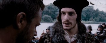 Movie still from “Robin Hood” (2010), directed by Ridley Scott – A man wearing a hat and a fur trimmed jacket talking to another man in front of a body of water; Close Up shot, Over the shoulder angle