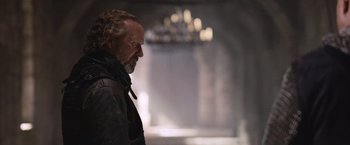 Movie still from “Robin Hood” (2010), directed by Ridley Scott – A man with a long beard standing in front of a chandelier; Close Up shot, Over the shoulder angle