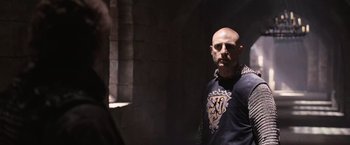 Movie still from “Robin Hood” (2010), directed by Ridley Scott – A bald man in a black t - shirt is standing in a dark room; Medium shot, Over the shoulder angle