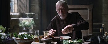 Movie still from “Robin Hood” (2010), directed by Ridley Scott – An older man sitting at a table with a plate of food in front of him; Medium shot, High angle