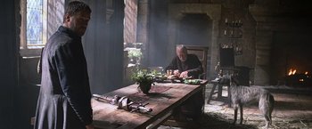 Movie still from “Robin Hood” (2010), directed by Ridley Scott – A man sitting at a wooden table in a room; Medium shot, Over the shoulder angle