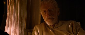 Movie still from “Robin Hood” (2010), directed by Ridley Scott – An older man sitting on a couch looking at the camera; Close Up shot, Over the shoulder angle