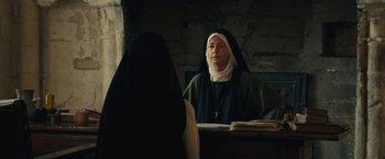 Movie still from “Benedetta” (2021), directed by Paul Verhoeven – A nun is sitting at a table in front of another nun; Medium shot, Low angle