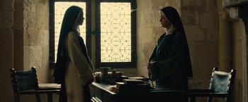 Movie still from “Benedetta” (2021), directed by Paul Verhoeven – A nun and a priest are standing in front of a window; Medium shot, Low angle