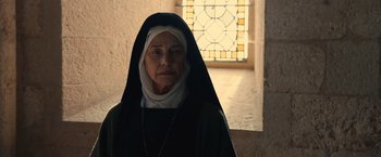 Movie still from “Benedetta” (2021), directed by Paul Verhoeven – An older woman wearing a nun's outfit in front of a stained glass window; Close Up shot, Low angle