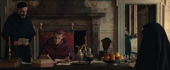 Movie still from “Benedetta” (2021), directed by Paul Verhoeven – A man sitting at a table in front of an open book; Medium shot, Low angle