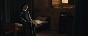Movie still from “Benedetta” (2021), directed by Paul Verhoeven – A man sitting on a chair next to a bed in a room; Wide shot, High angle
