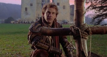 Movie still from “Robin Hood: Prince of Thieves” (1991), directed by Kevin Reynolds – A man in medieval garb sitting next to a tree; Medium shot, Over the shoulder angle
