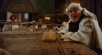 Movie still from “Robin Hood: Prince of Thieves” (1991), directed by Kevin Reynolds – An older man sitting at a table with a bag; Medium shot, High angle