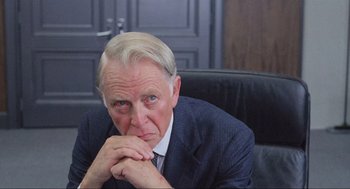 Movie still from “RoboCop” (1987), directed by Paul Verhoeven – An older man sitting in a chair with his hands folded to his chest; Close Up shot, Over the shoulder angle