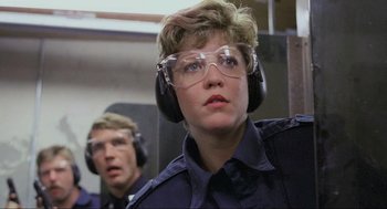 Movie still from “RoboCop” (1987), directed by Paul Verhoeven – A woman wearing ear muffs and glasses while looking at the camera; Close Up shot, Low angle