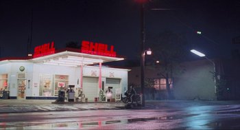 Movie still from “RoboCop” (1987), directed by Paul Verhoeven – A man sitting on the side of the road next to a gas station; Extreme Wide shot, Low angle