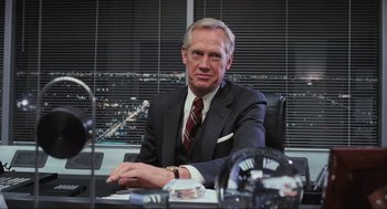 Movie still from “RoboCop” (1987), directed by Paul Verhoeven – An older man sitting at a desk in a suit and tie; Medium shot, Over the shoulder angle