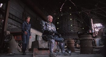Movie still from “RoboCop” (1987), directed by Paul Verhoeven – A man and a woman standing next to each other in front of a robot; Wide shot, Low angle