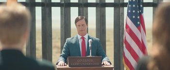 Movie still from “Robots” (2023), directed by Casper Christensen – A man wearing a suit and red neck tie is giving a speech; Medium shot, Over the shoulder angle