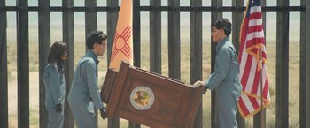 Movie still from “Robots” (2023), directed by Casper Christensen – Two men carrying a wooden podium in front of a fence; Wide shot, Over the shoulder angle