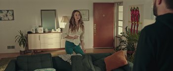 Movie still from “Robots” (2023), directed by Casper Christensen – A woman standing in a living room next to a couch; Wide shot, Over the shoulder angle