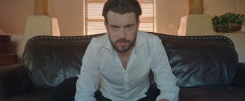 Movie still from “Robots” (2023), directed by Casper Christensen – A man sitting on top of a couch wearing a white shirt; Medium shot, Low angle