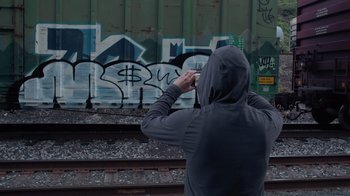 Movie still from “Rolling Like Thunder” (2021), directed by Roger Gastman – A man in a hooded sweatshirt is standing on train tracks; Wide shot, Low angle