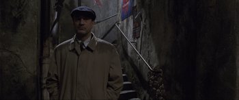 Movie still from “Ronin” (1998), directed by John Frankenheimer – A man in a trench coat standing in front of stairs; Medium shot, Low angle