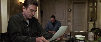 Movie still from “Ronin” (1998), directed by John Frankenheimer – A man sitting at a table looking at papers; Medium shot, Low angle