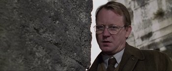 Movie still from “Ronin” (1998), directed by John Frankenheimer – A man in a suit and tie standing next to a stone wall; Close Up shot, Low angle