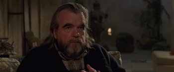Movie still from “Ronin” (1998), directed by John Frankenheimer – An older man with long hair and a goatee is sitting on a couch; Close Up shot, Over the shoulder angle