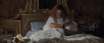 Movie still from “Rosaline” (2022), directed by Karen Maine – A woman sitting on a bed reading a book; Medium shot, High angle