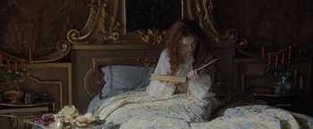 Movie still from “Rosaline” (2022), directed by Karen Maine – A woman sitting on a bed reading a book; Medium shot, High angle