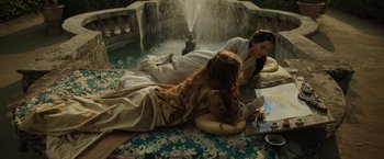 Movie still from “Rosaline” (2022), directed by Karen Maine – Two women laying on the ground in front of a fountain; Medium shot, High angle