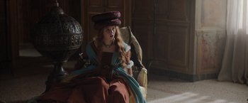 Movie still from “Rosaline” (2022), directed by Karen Maine – A woman sitting in a chair wearing a hat; Medium shot, Over the shoulder angle
