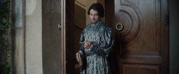 Movie still from “Rosaline” (2022), directed by Karen Maine – A man standing in front of a door holding a book; Medium shot, Low angle