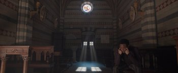 Movie still from “Rosaline” (2022), directed by Karen Maine – A man sitting in a church with a stained glass window; Wide shot, Low angle