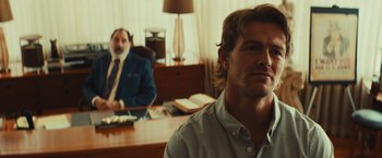 Movie still from “Run & Gun” (2022), directed by Christopher Borrelli – A man sitting at a desk in front of another man in a suit; Close Up shot, Over the shoulder angle
