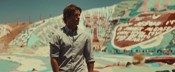 Movie still from “Run & Gun” (2022), directed by Christopher Borrelli – A man standing in front of a wall with graffiti on it; Medium shot, Low angle